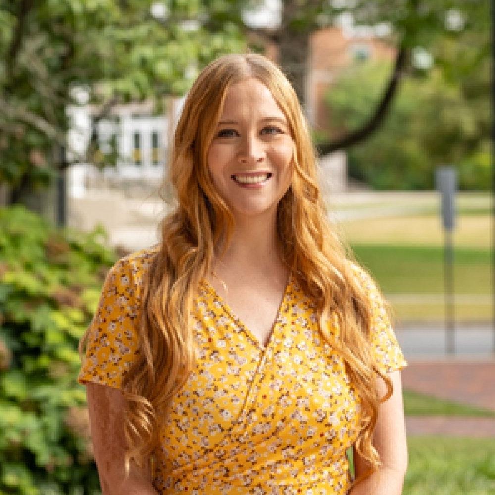 Lady with blonde hair wearing yellow floral dress outside on campus